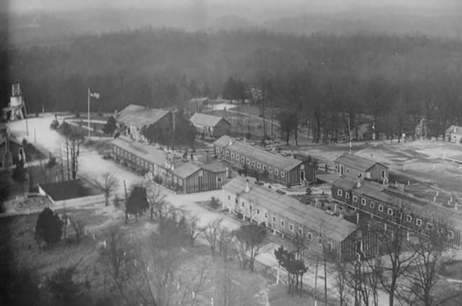 A black and white aerial view of a camp with long, one-story buildings, a network of roads, a water tank, and other features.