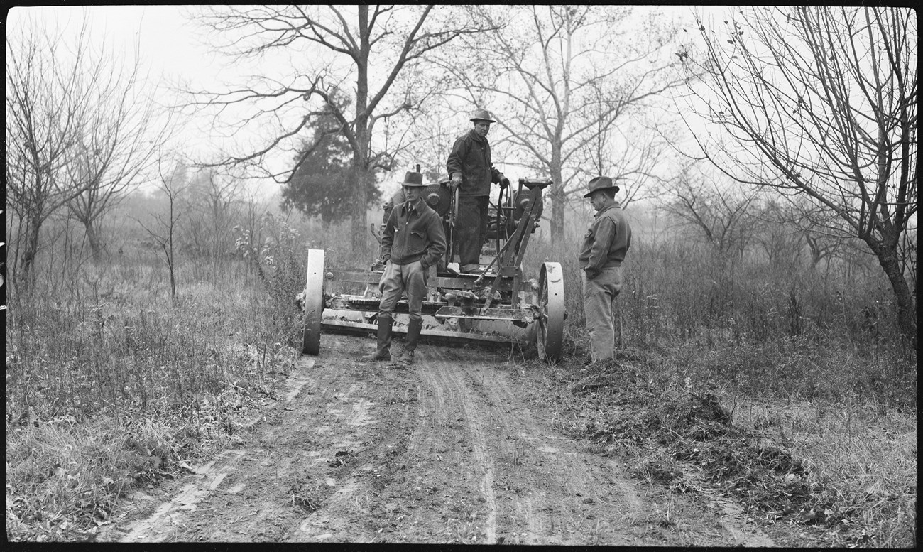 Black and white photo of three men in a landscape of leafless trees and unpaved road. Two stand in the roadbed, and one stands on the platform of a piece of machinery used to grade the road.