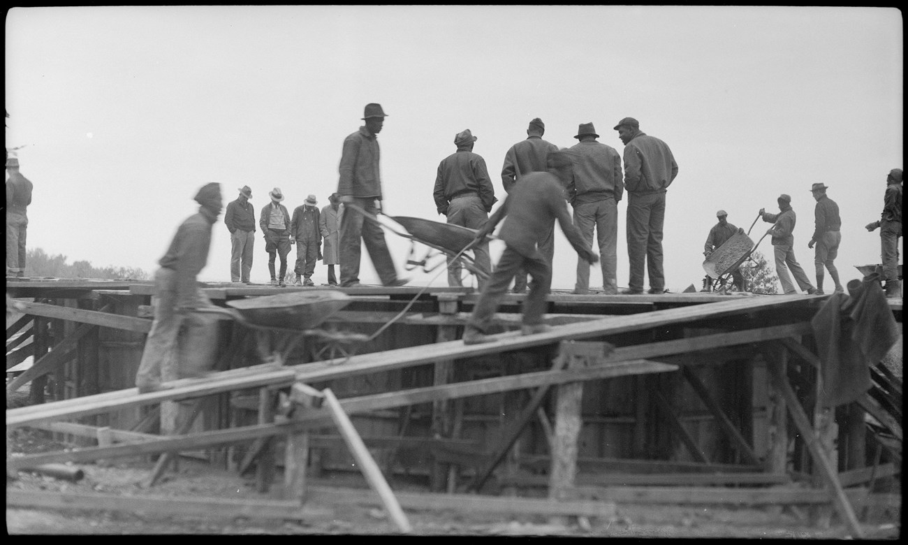 Black and white photo of a group of men working on construction. Two people push wheelbarrows up a wooden ramp.