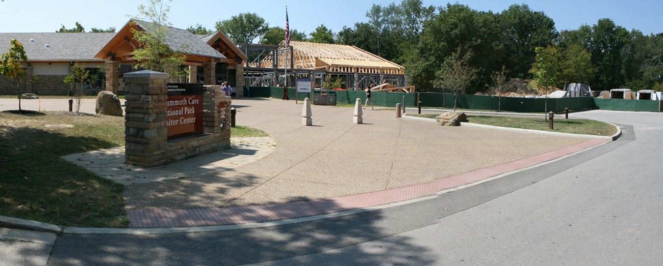 New building construction is visible behind an existing stone, one-story visitor center with a concrete patio connecting it to the road.