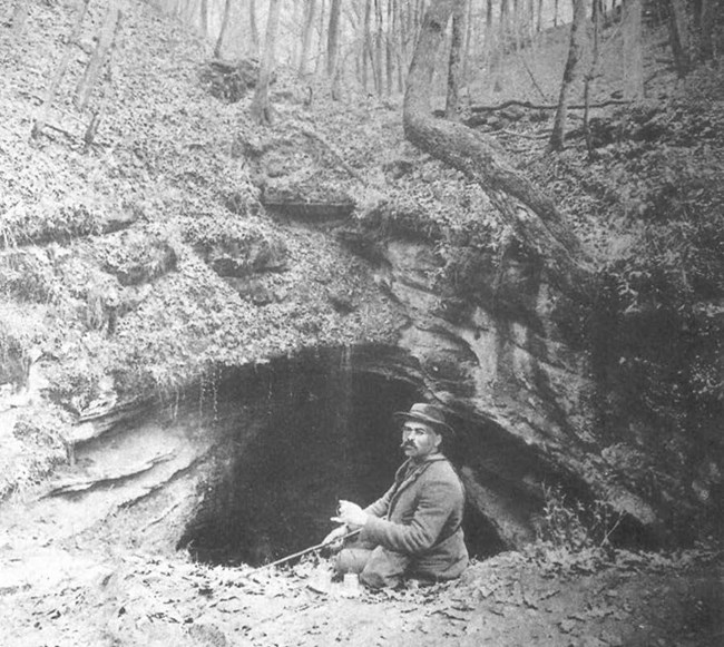 A man in a suit sits on the ground in front of the opening to a cave, surrounded by forest.