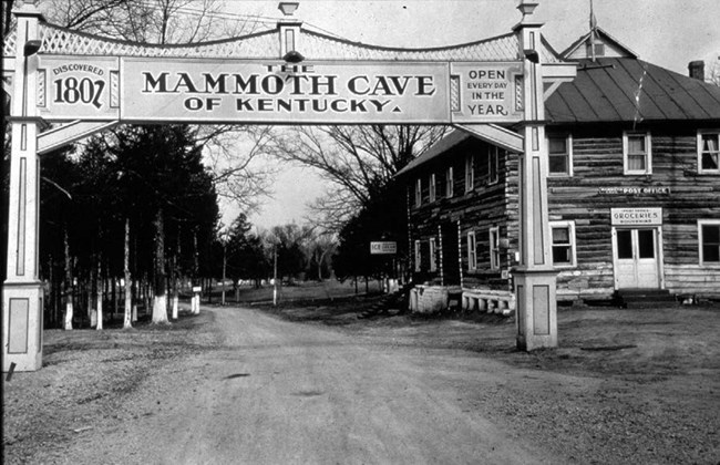 An archway entrance over an unpaved road, with two square pillars holding a sign "The Mammoth Cave of Kentucky, Discovered 1802, Open Every Day In The Year." A wooden building with groceries and post office signs is to the right of the road.