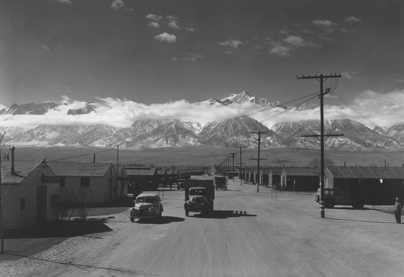 Trucks drive down a wide, dusty road between rows of barracks, with mountains in the background