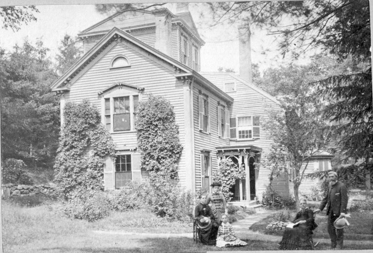 Five people, some seated and some standing, in a yard beside a house framed by vines and several trees in the 1880s