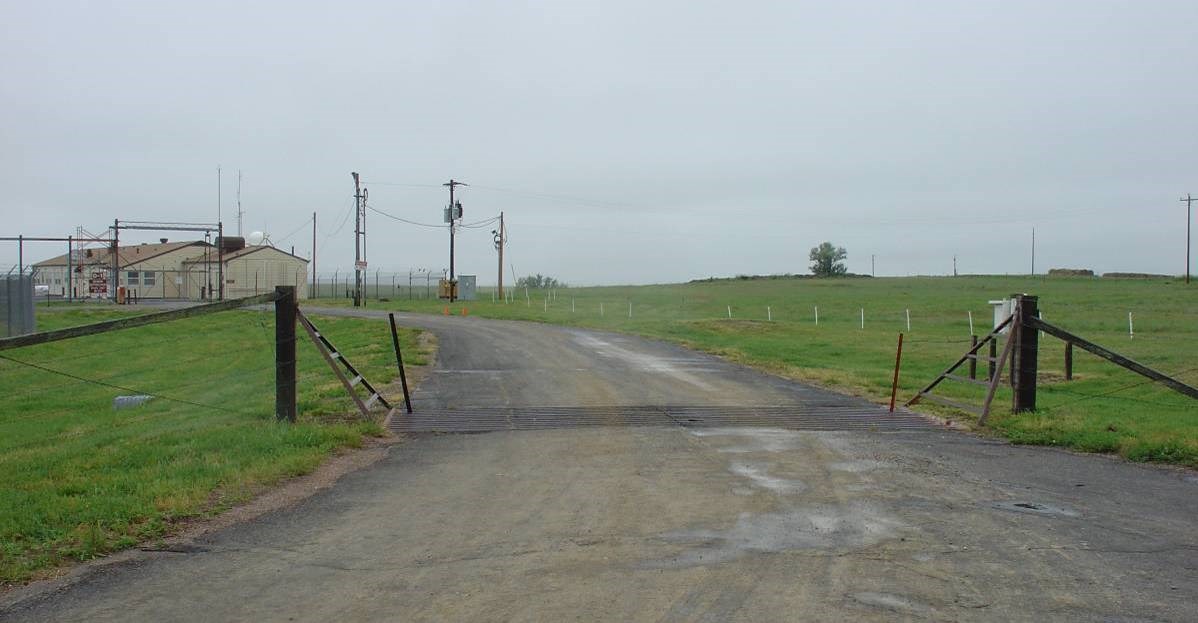 A road through a gate towards low buildings, surrounded by fences