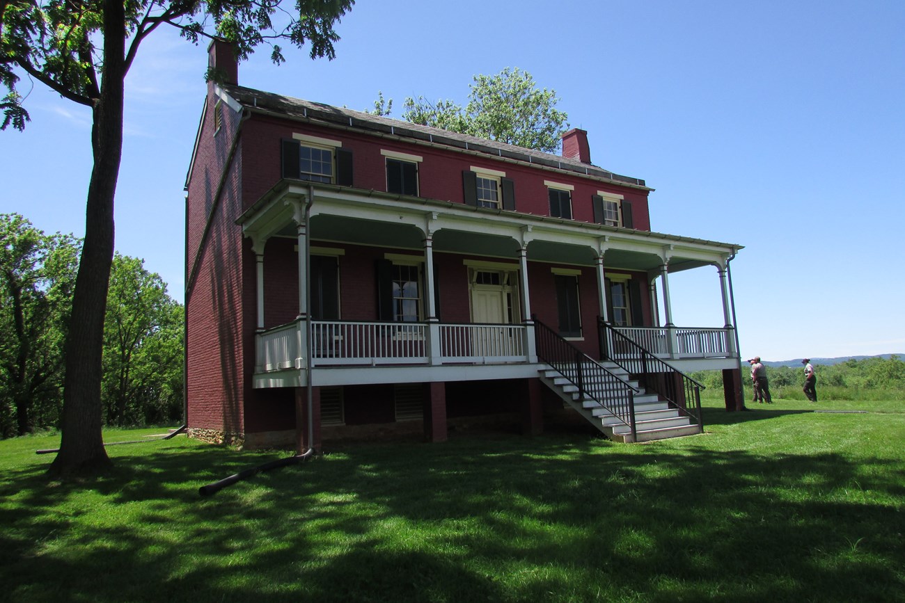 A two-story brick farmhouse with a front porch acrosss the front, beside a treeline and open turf