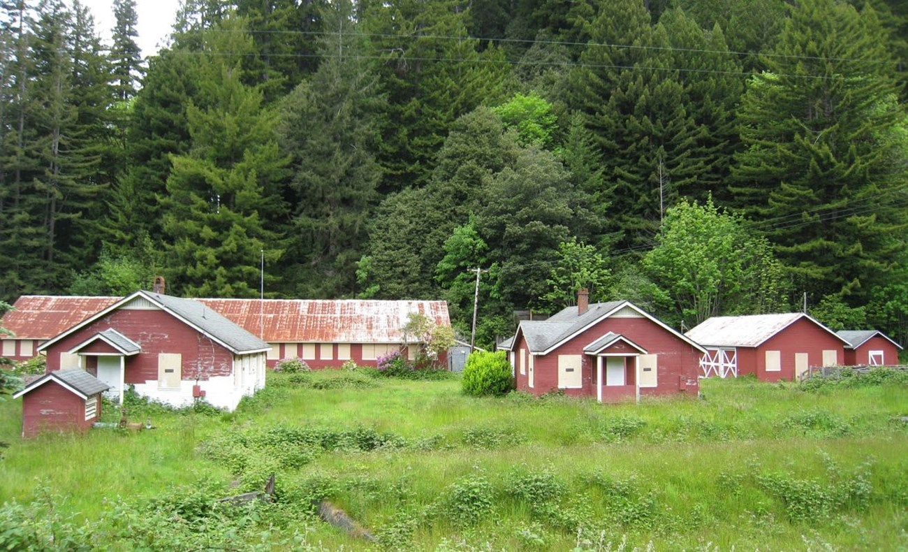 One-story wooden buildings, red with white trim, are clustered in a level grassy area with a tree-covered hillside in the background