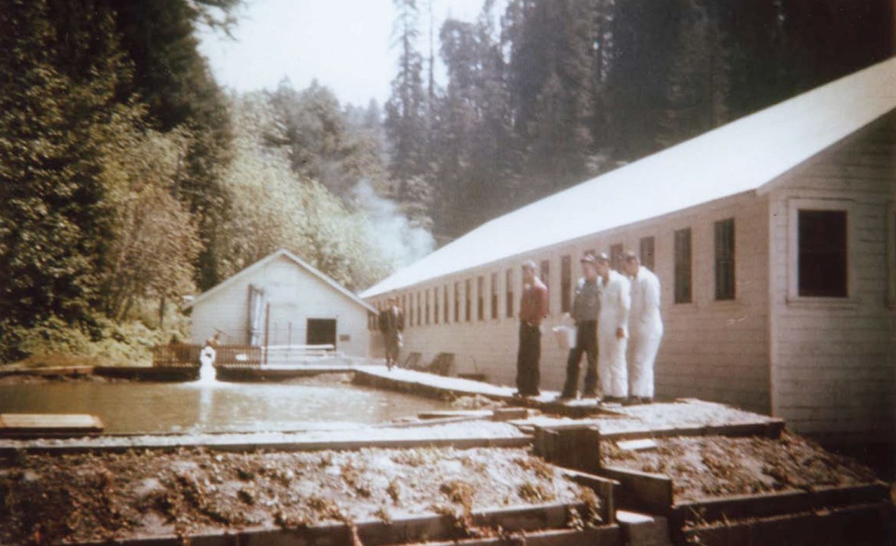 A group  of people stand on a walking platform that surrounds an aboveground earthen pond beside Hatchery Building