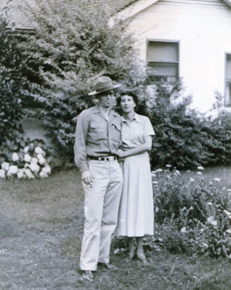 Two people stand in a yard in front of the Assistant Superintendent’s House, near a garden bed, foundation shrubs, and turf.
