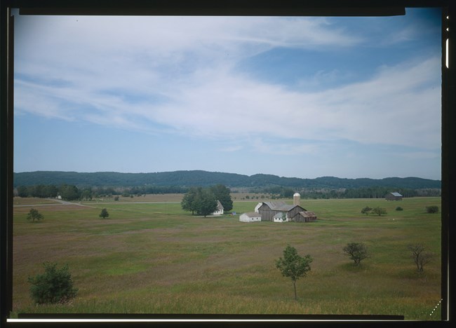 A cluster of farm buildings, including a house in a group of trees, stands in the middle of an open field.