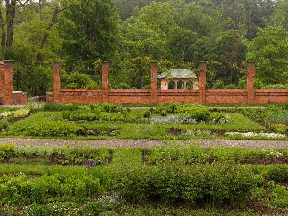 A formal garden surrounded by a brick wall, contains rectangular planting beds, turf, and walkways.