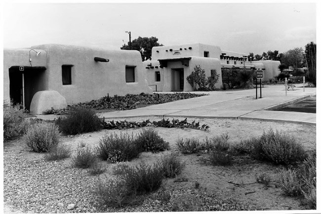 Comfort station at left and Administration and Museum Building at right. In front of the rounded adobe buildings, there is a concrete walkway and scrubby vegetation.