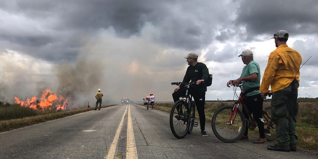 Bicyclists pass by a prescribed fire. Bicylclist on the road next to a prescribed fire stop to talk to firefighters.