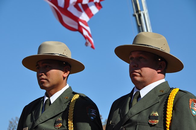 Two men in dress NPS uniform stand beneath a United States flag.