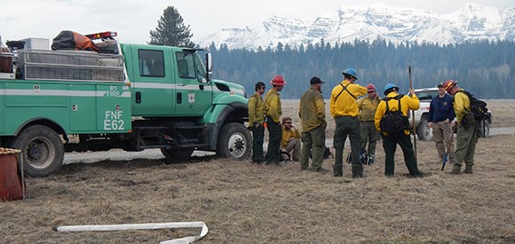 A fire crew stands near an engine with snowy mountains in the distance.