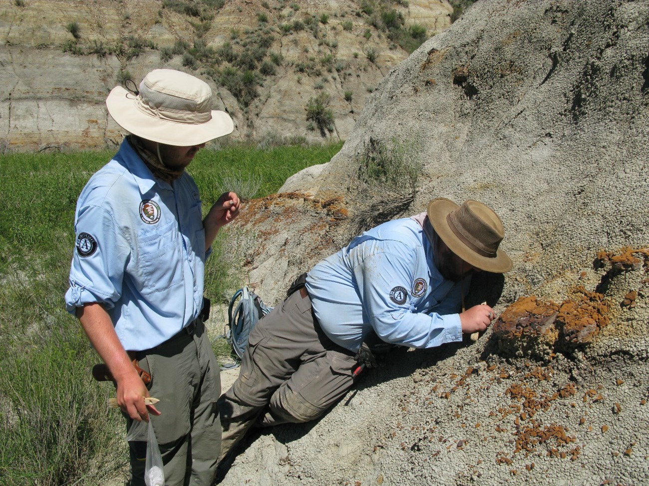 THRO_paleontology_inventory_2022_photo 2 A photo of two men in uniforms examining the rounded base of a bluff of soft sedimentary rock.