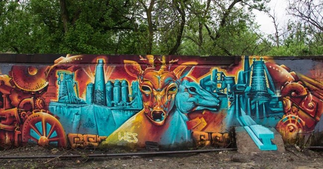 An orange deer head and a blue deer head sit in front of blue iron works. A mural.
