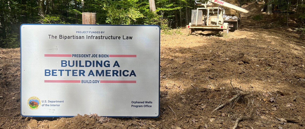 a white sign sits in a tree lined dirt lot, the sign reads "Project Funded by The Bipartisan Infrastructure Law - Building a Better America"