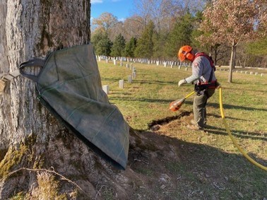 An NPS employee wearing safety gear uses an air powered excavation spade to form a trench extending out from the base of a tall tree