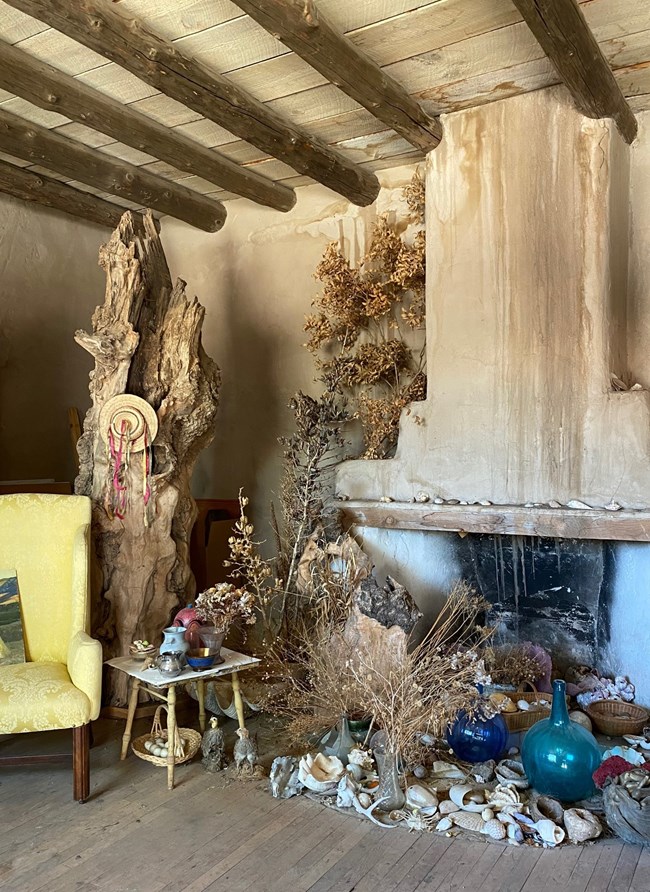 Arrangement of shells, bottles, pitchers, taxidermized birds, dried flowers, baskets, a small table, and other objets d'art sits on the floor in front of a large adobe fireplace. Part of a tree trunk adorned with a hat with ribbons is at left behind a yel