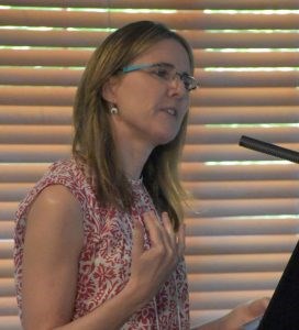 White woman with long sandy brown hair and glasses wearing a sleeveless res and white leaf pattern top. Standing at a microphone.