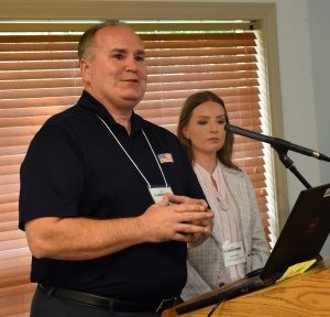 A white male wearing a black polo shirt stands at a podium and a white woman in a pale gray jacket stands to his left.