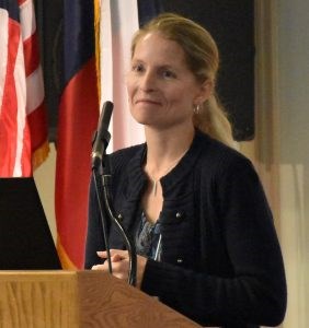 White woman with a blond ponytail wearing a black sweater standing at a podium.