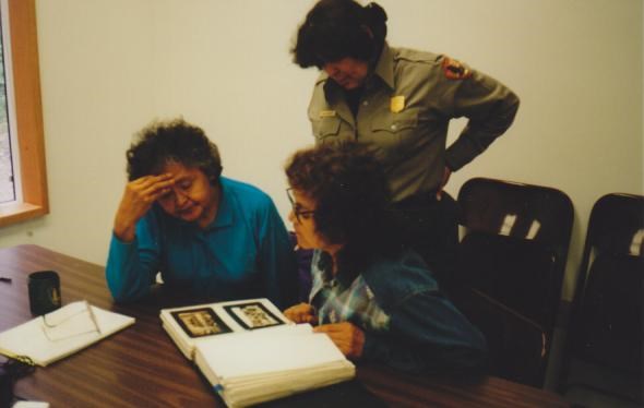 Two women are seated at a table looking at a photo album, as a woman in NPS uniform looks over their shoulder