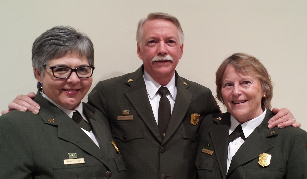 Three people, Kathleen Bond, Jon Jarvis, and Laura Gates, pose in the green jacket and pins of NPS uniform,