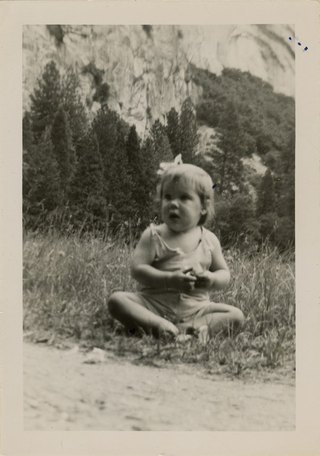 Laurel Boyers, as a young child, sits cross-legged on the ground with trees and mountains in the background.