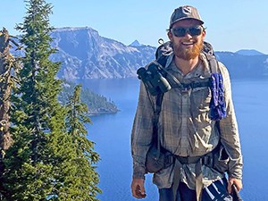 Long-Distance Hikers Navigate the Hazards of a Changing Climate a smiling man with a red beard wearing a backpack stands in front of a blue lake with mountains in the distance