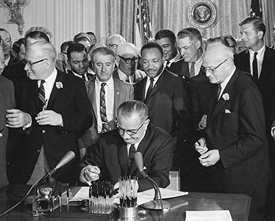 photo of Lyndon Johnson signing a document with Martin Luther King Jr and others standing behind him