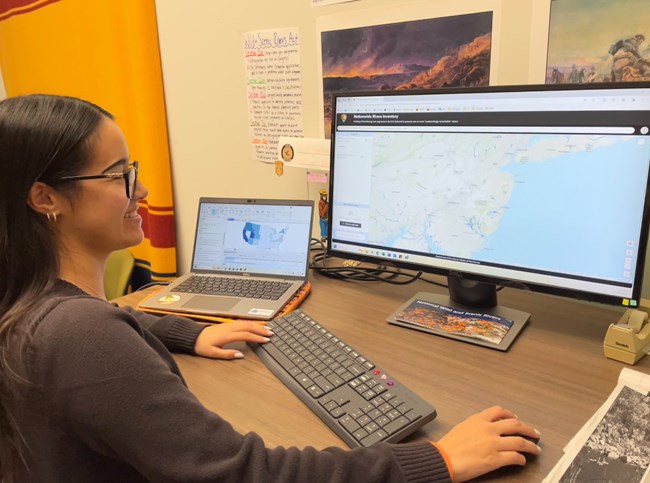 Nationwide Rivers Inventory GIS Fellow A young woman smiles and stands at her desk in front of a computer with the 2024 NRI data viewer on the screen.