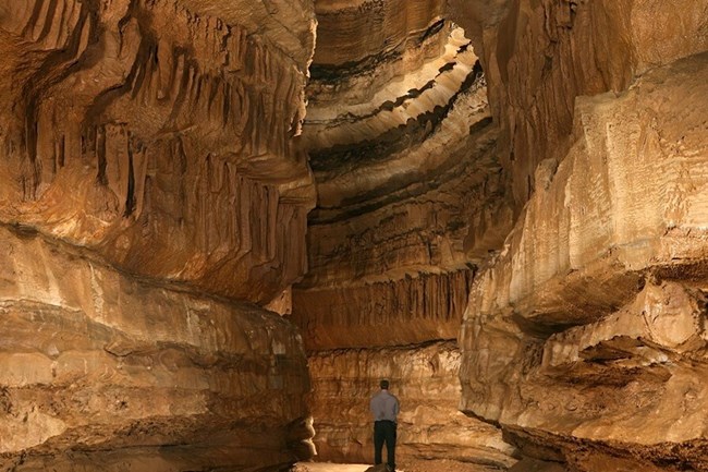 A park ranger is dwarfed by the reddish rocks of a large cave.