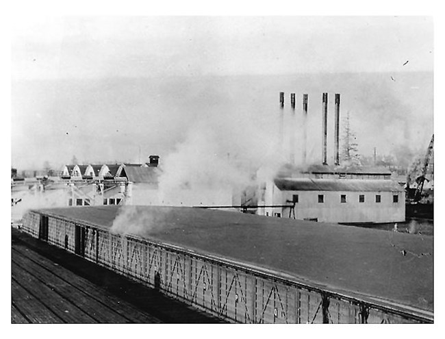 A black and white photo of steam rising from the long, wooden dry kiln building.