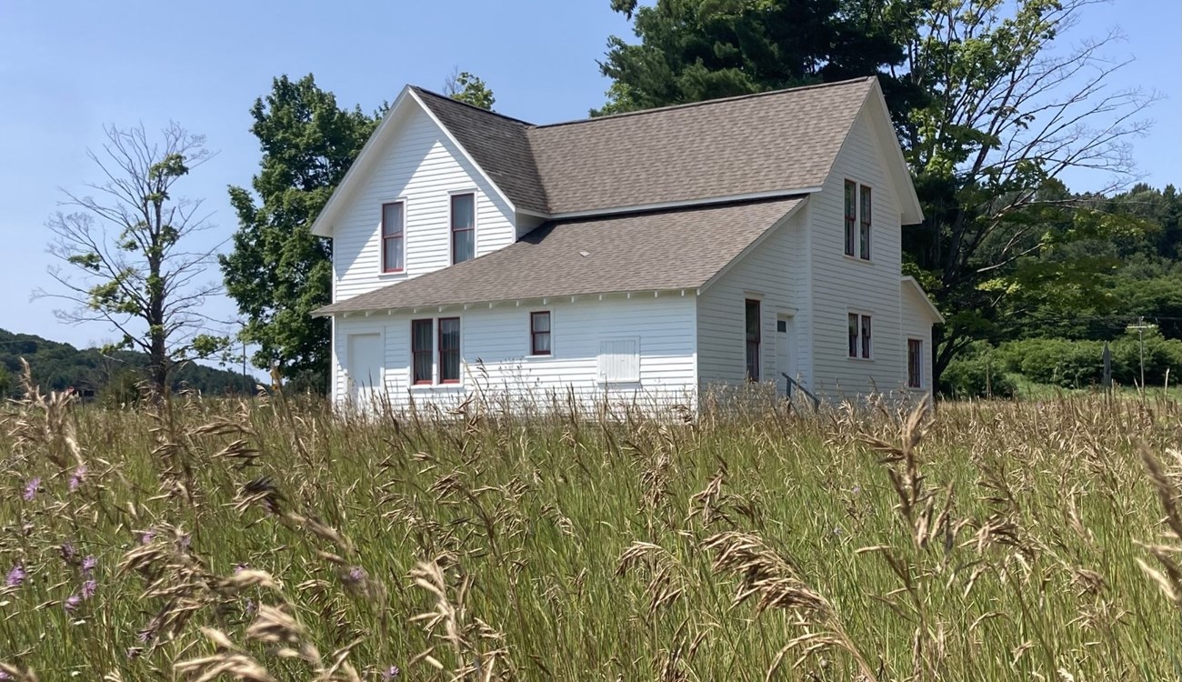 Kelderhouse2024-StotzGhosh-crop Amongst seeding green grass, a white farmhouse with red trimmed windows, a bright blue sky and several trees poking out from behind the house.