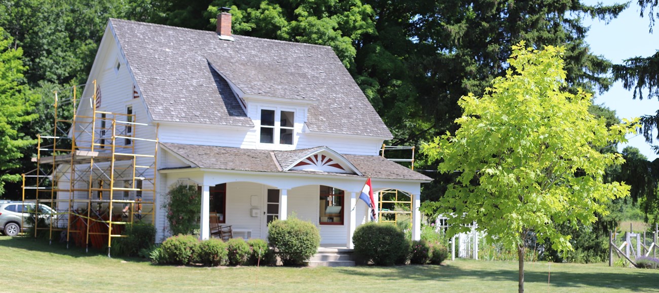 Olsen's white two-story farmhouse basks in the sun, red details of the porch overhang, and the red, blue, and white "open" flag greet you. Sides of the house with yellow scaffolding for a project, while the front porch is surrounded by green circular trim