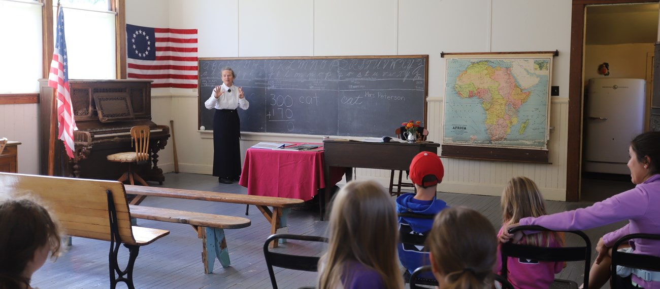 Inside of a historic one-room schoolhouse, surrounded by a piano, chalkboard with the alphabet, and a map of Africa, Mrs. Peterson dressed in a long black skirt and white button-up shirt teaches today's youth.