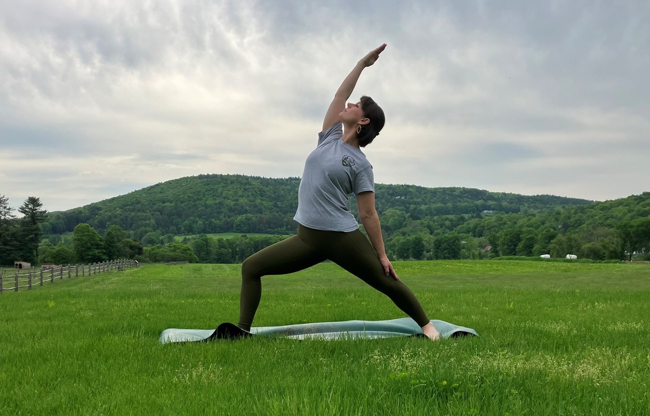 person in gray National Park Service t shirt does a yoga pose on a yoga mat with a green hillside and farm in the background