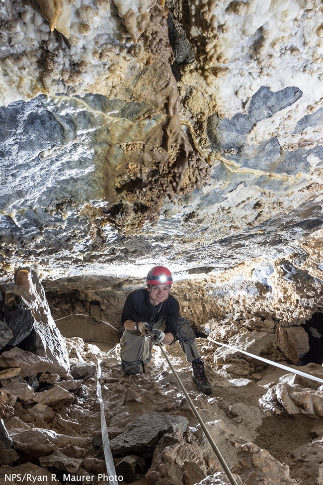 Caver sits smiling while holds onto a rope handline in Hansen Cave, surrounded by rocks