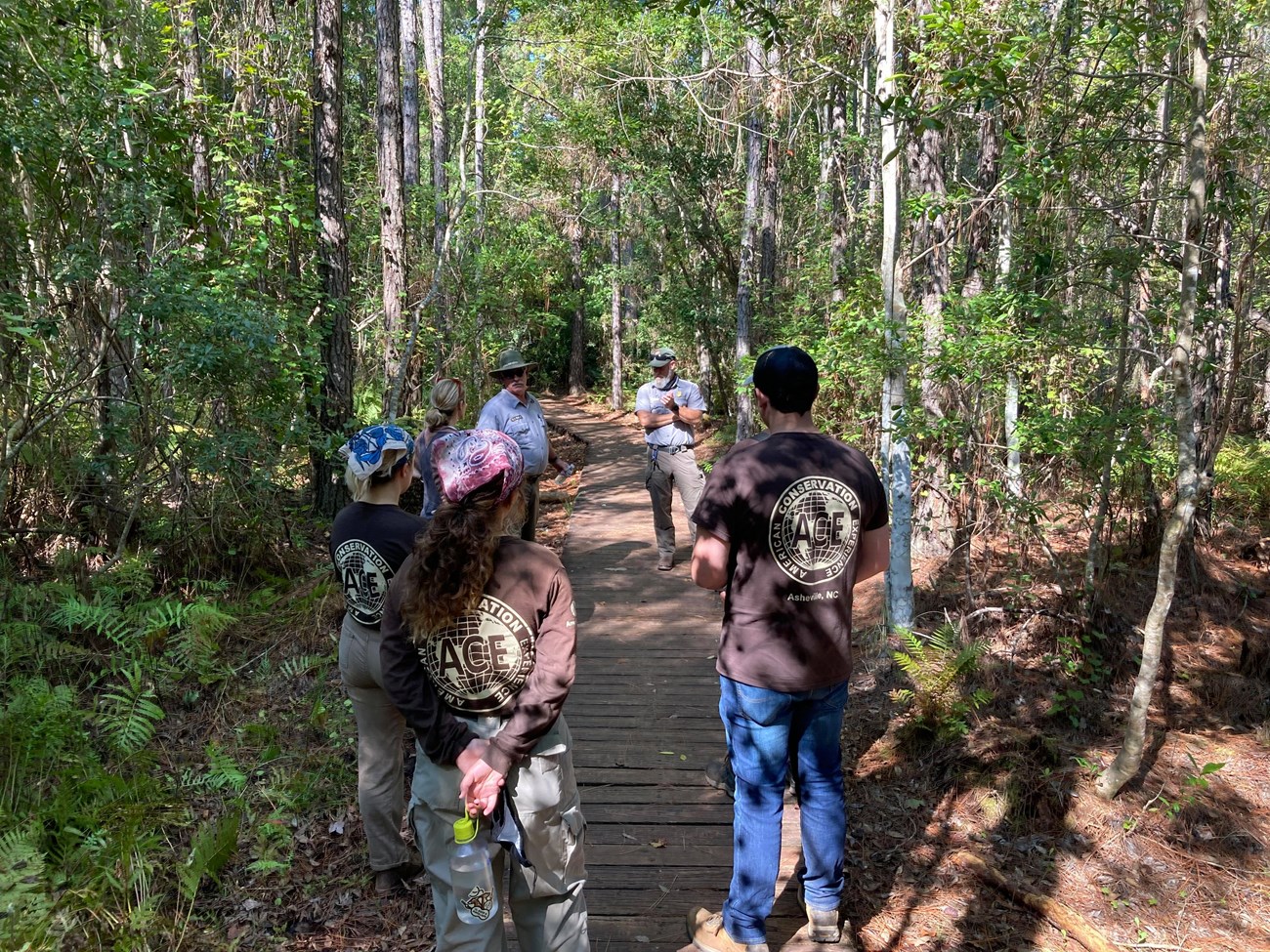 youth in brown ACE shirts talk to rangers on boardwalk in the woods