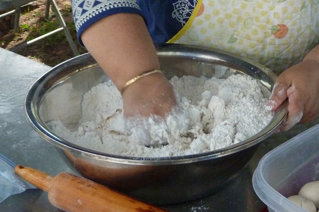 Hands mixing flour, salt, lard, and water in a metal mixing bowl.