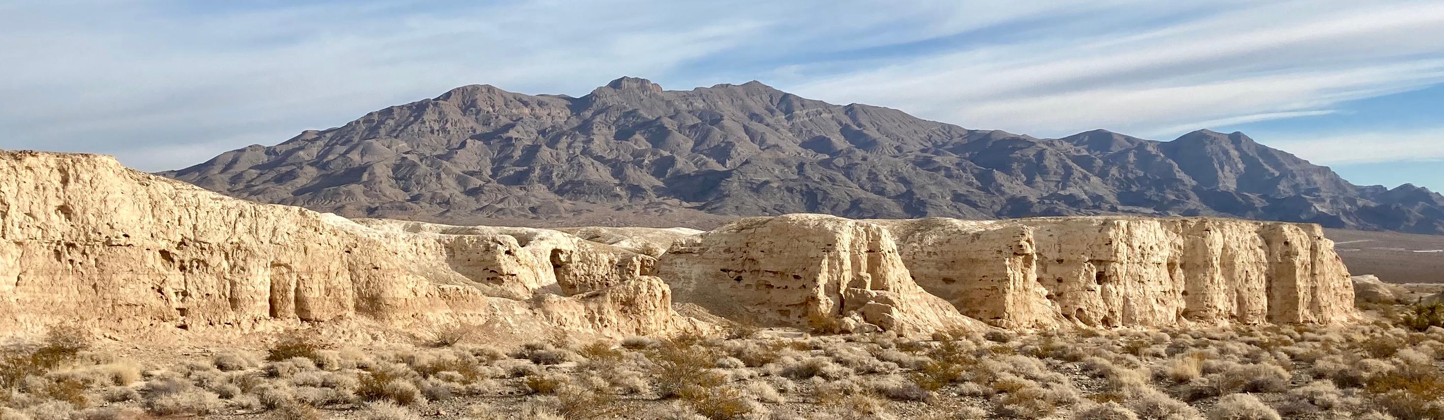 Sunlit badlands in a desert environment with a darker limestone mountain range in the background