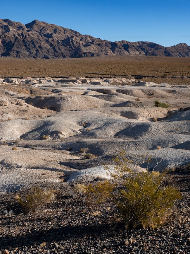 Tan badlands emerge below a mountain range in a desert landscape.