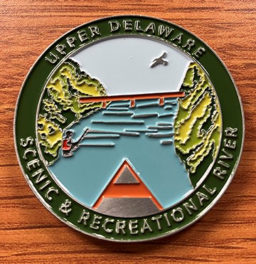 metal medallion with graphic of bridge over a river connecting 2 hillsides. Canoe & fisherman in river. Eagle in sky. Text: Upper Delaware Scenic & Recreational River.