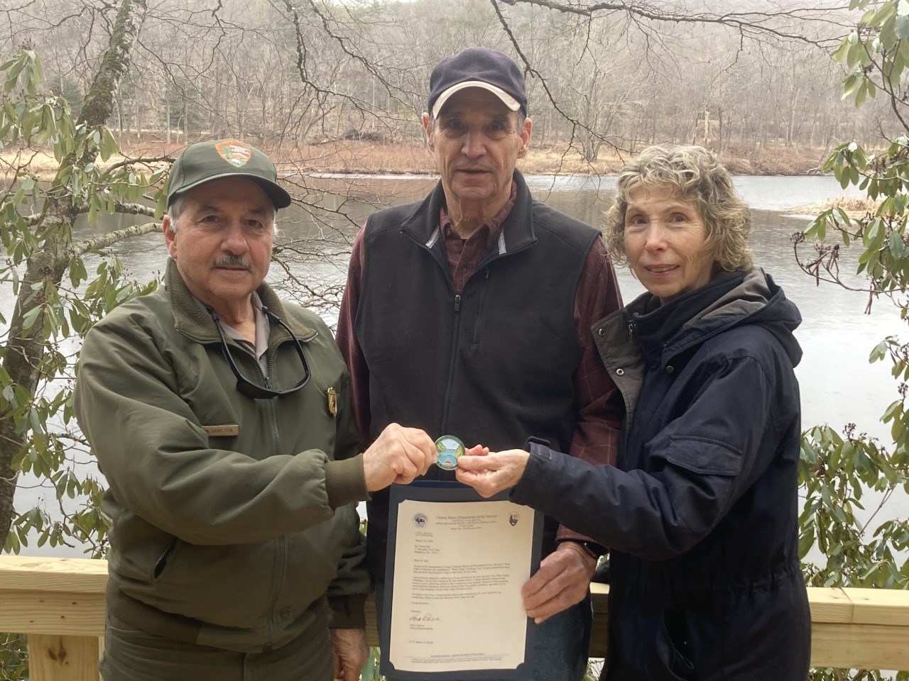 three people standing together. On left is park ranger wearing green. In center is tall man in ball cap and vest. On right is woman with blond, curly hair. Man on left and woman hold a large decorative medallion in the center. Man in center holds letter.