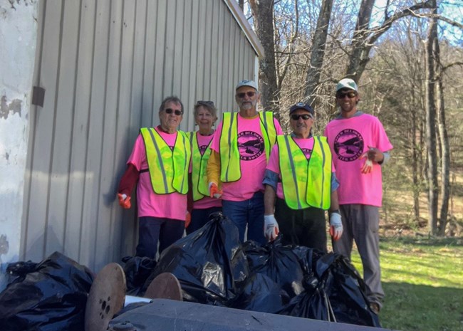 Group of five men in UDC litter sweep T-shirts and safety vests smile at camera with large bags of trash.