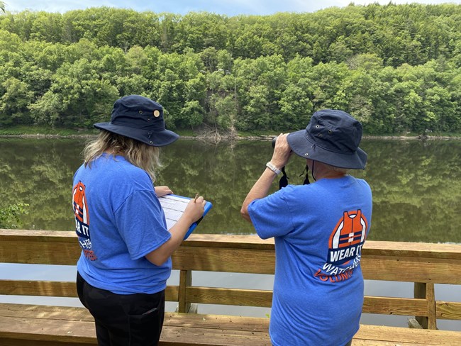 Two volunteers standing against wooden deck, looking out onto river with binoculars