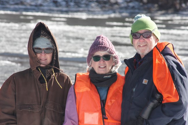 Three volunteers in warm hats and bright orange vests smile at camera