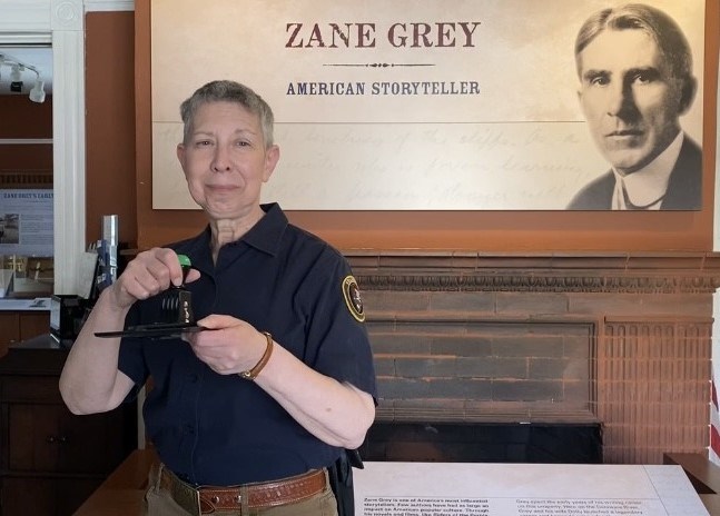 Volunteer stands in front of sign of Zane Grey, holding passport stamp